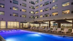 A hotel pool area with lounge chairs and umbrellas, surrounded by a multi-story building with illuminated windows.