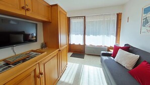 A compact living space with wooden cabinets, a flat-screen TV, a sofa with red cushions, and a window with sheer curtains. - 2-room apartment, sleeps 6, terrace, garage, Le Grand-Bornand (Le Grand-Bornand)