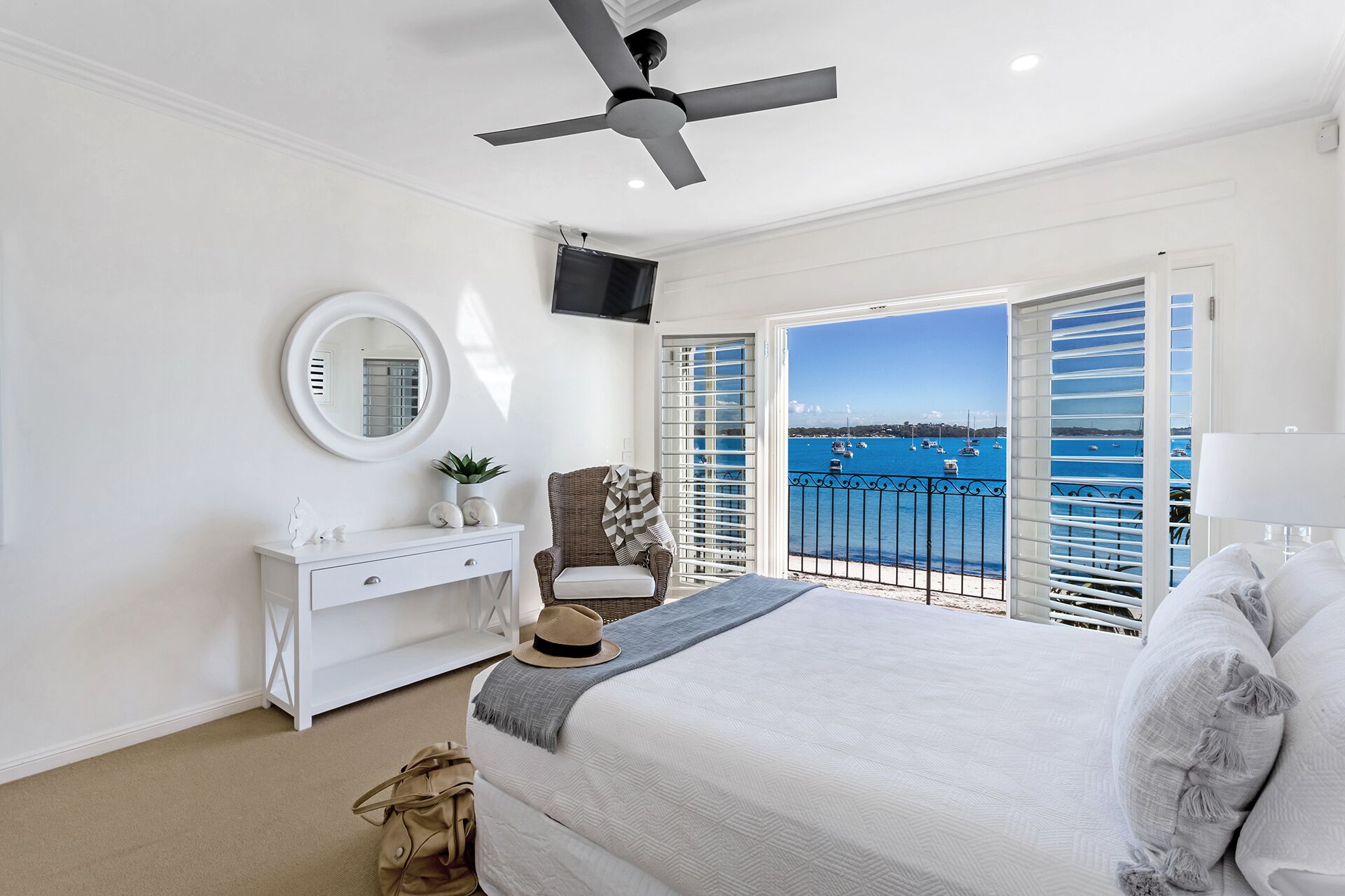 A bedroom with a bed, a dresser, a mirror, a ceiling fan, and a view of the sea through open shutters.