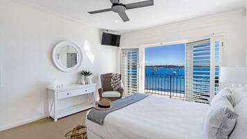 A bedroom with a bed, a dresser, a mirror, a ceiling fan, and a view of the sea through open shutters.