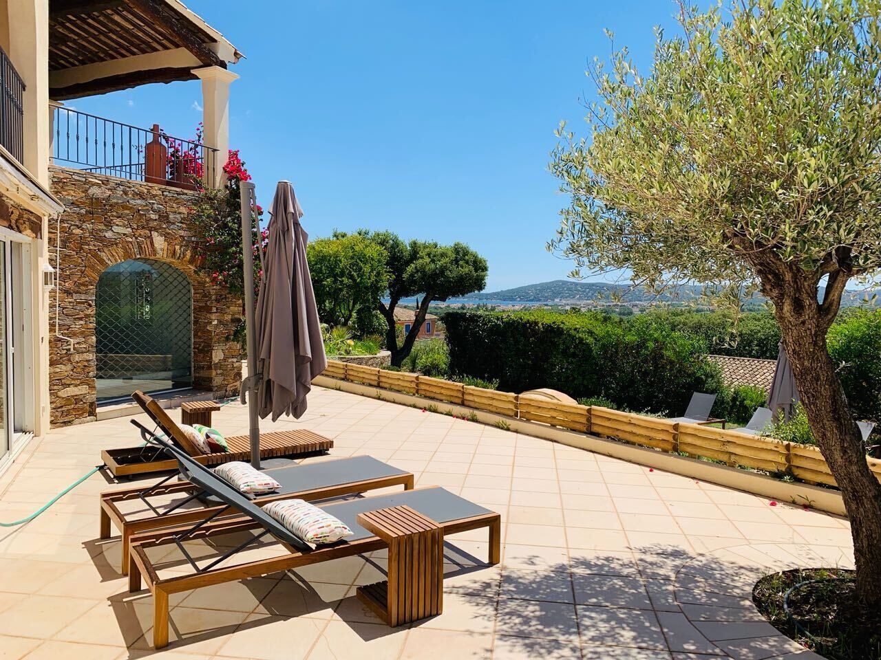 A sunlit outdoor seating area with lounge chairs and a covered umbrella, overlooking a scenic landscape with trees and distant hills.