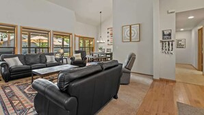A spacious living room with a black leather sofa set, a coffee table, and a dining area with a table and chairs. Large windows provide natural light and views of the outdoors. - AC, Hot Tub, One level, Pet friendly (Sunriver)