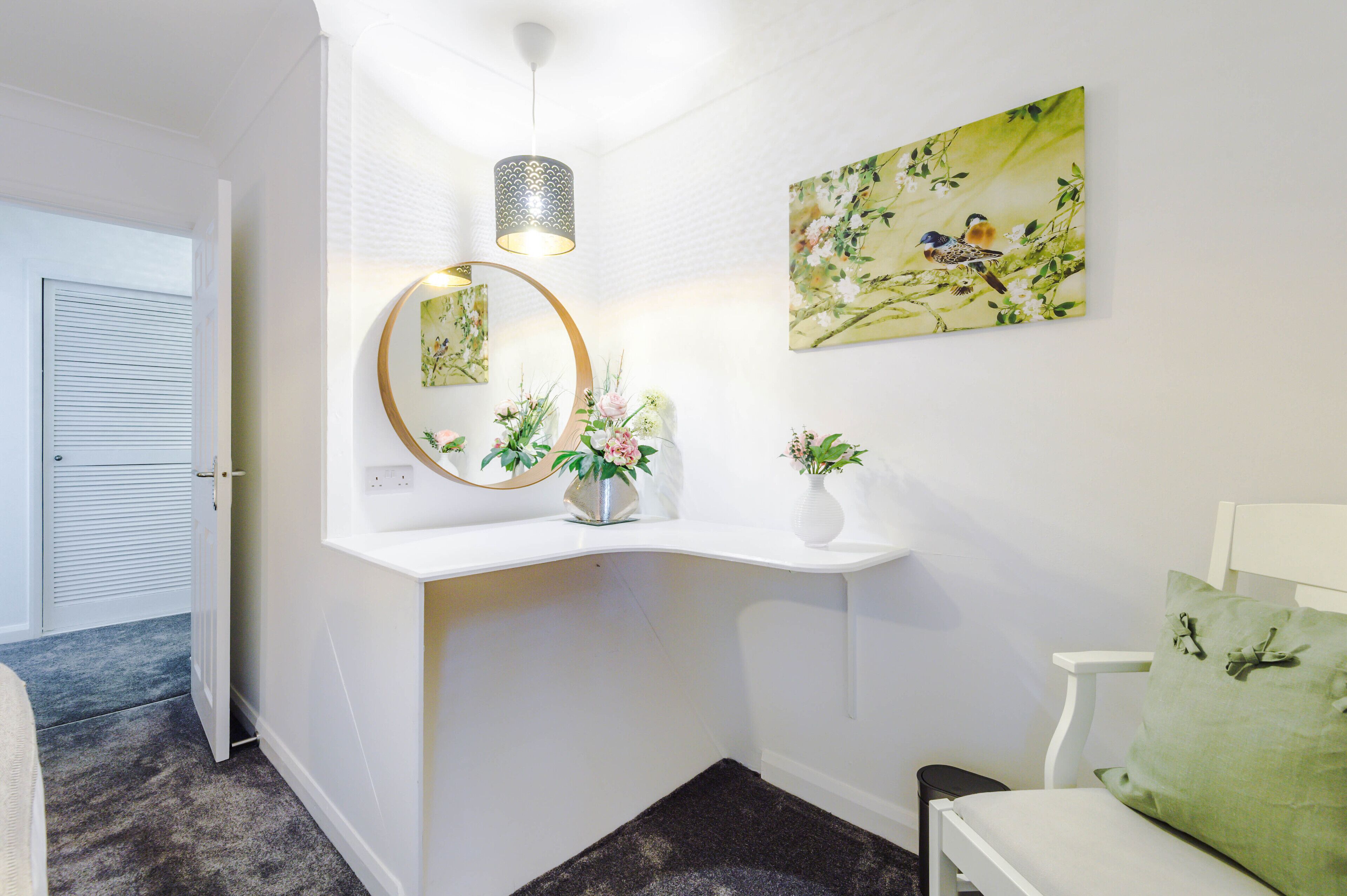 Een gang met een witte muur, een ronde spiegel, een vaas met bloemen, een schilderij van vogels en een groen kussen op een witte stoel.