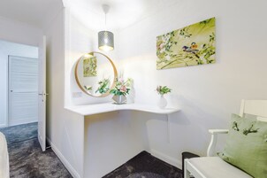 A hallway with a white wall, a round mirror, a vase with flowers, a painting of birds, and a green pillow on a white chair.