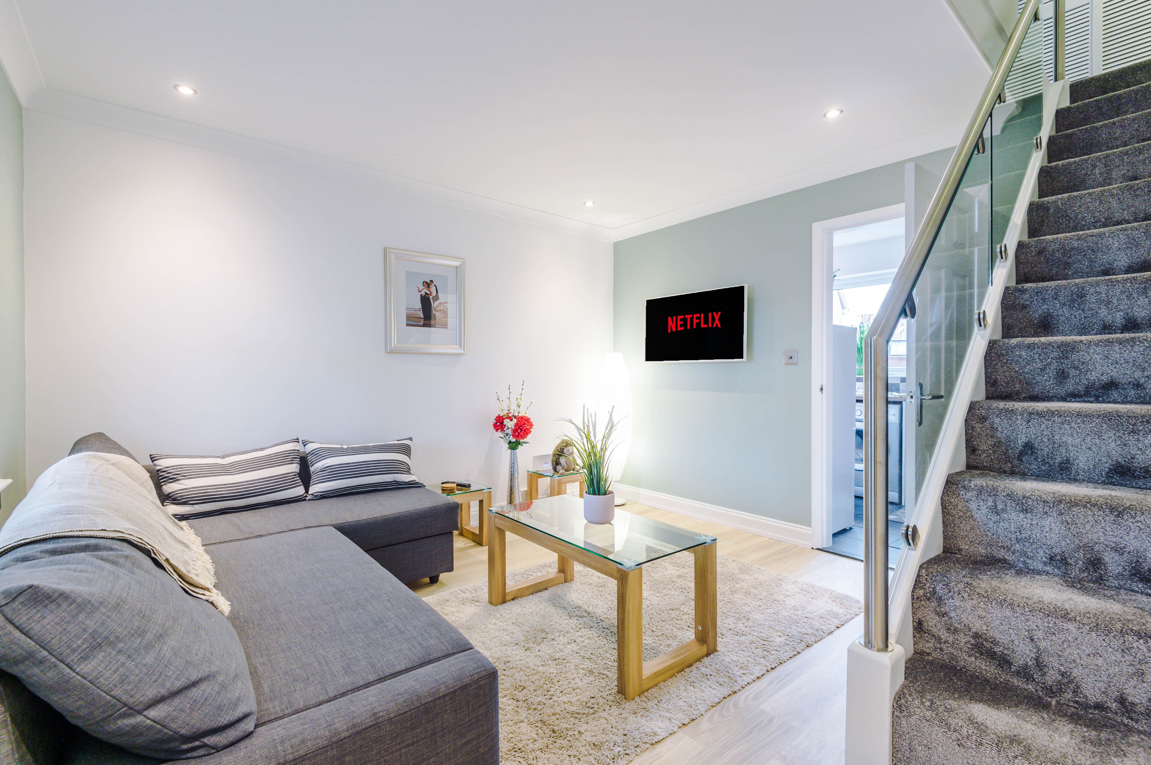 A modern living room with a grey sofa, a glass coffee table, and a staircase with a glass railing.