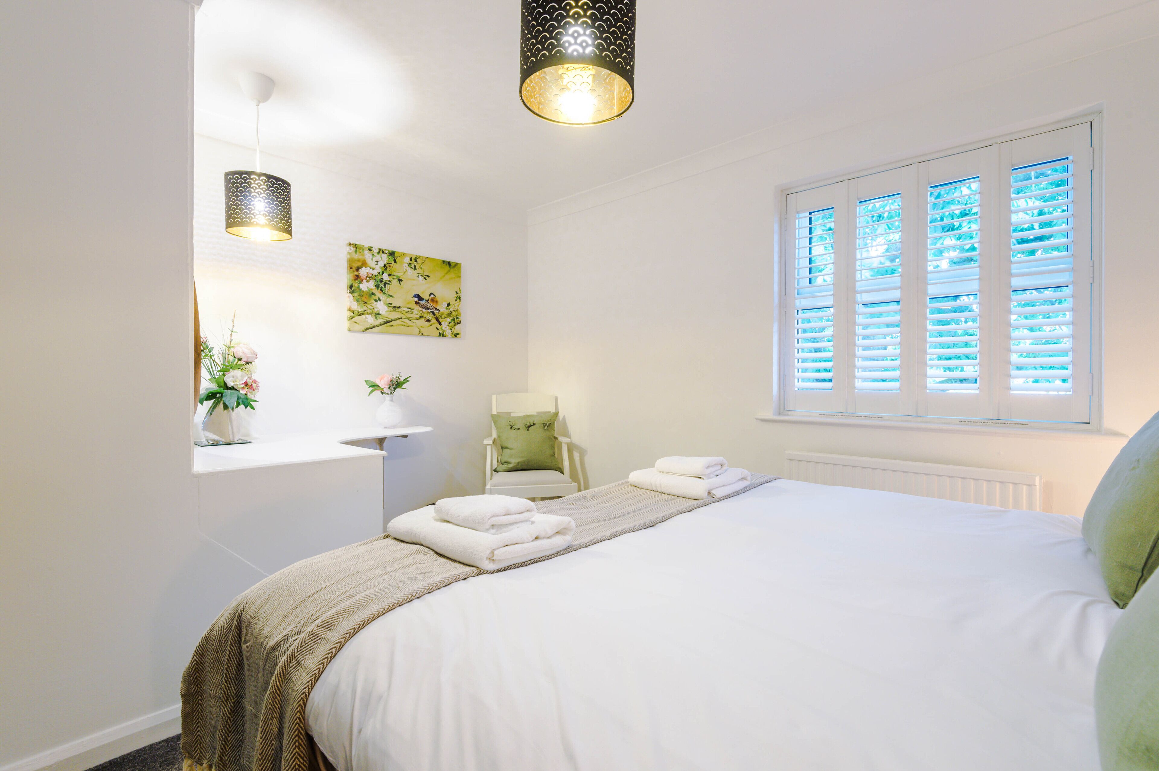 A bedroom with a bed, a window with blinds, a chair, a bedside table, and a wall-mounted light fixture.