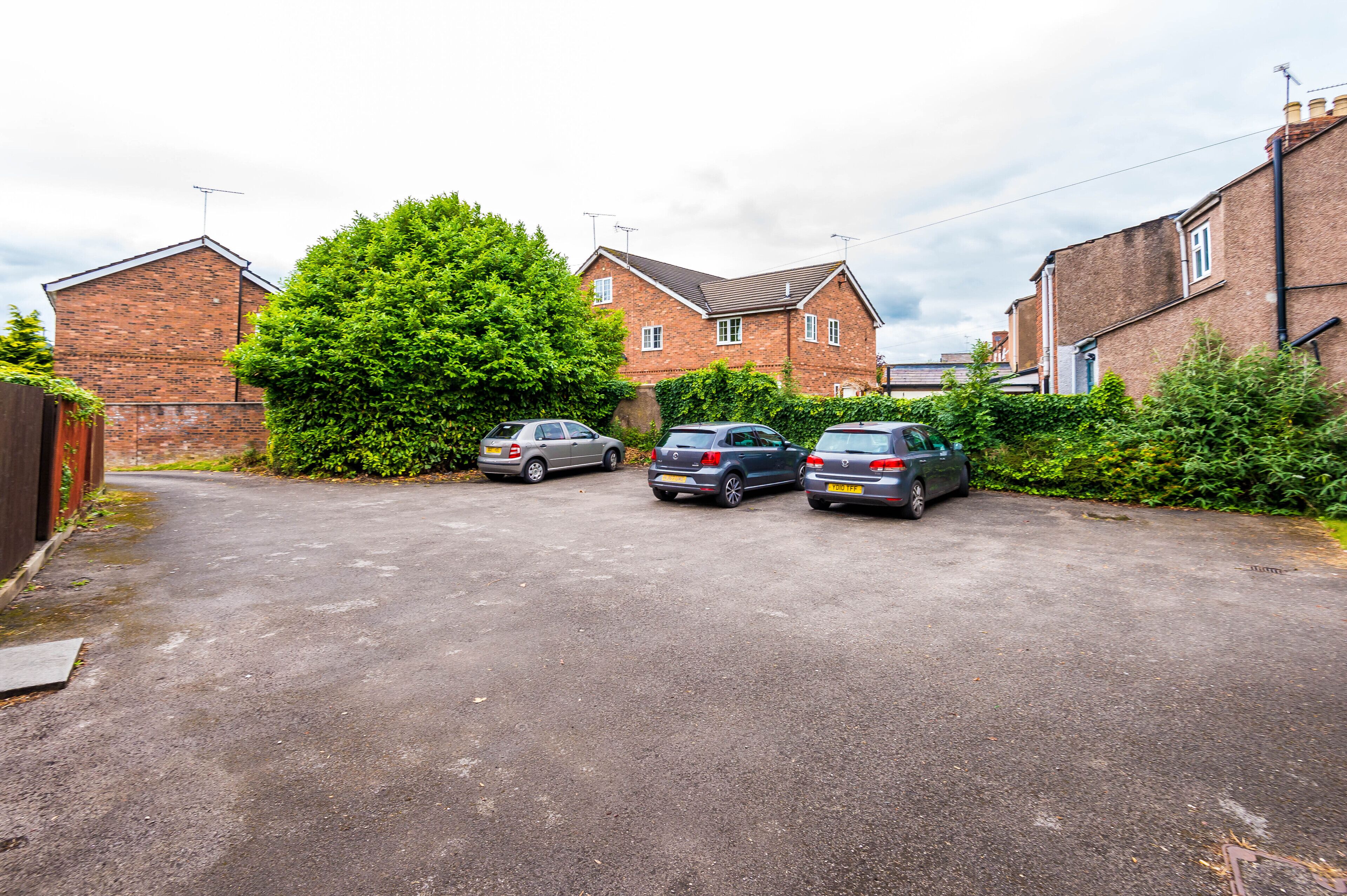 A residential street with three parked cars, brick houses, and a large tree.