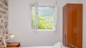 Ein Schlafzimmer mit einem Bett, einem Nachttisch mit Lampe, einem Holzschrank und einem Fenster mit Blick auf Grünflächen.