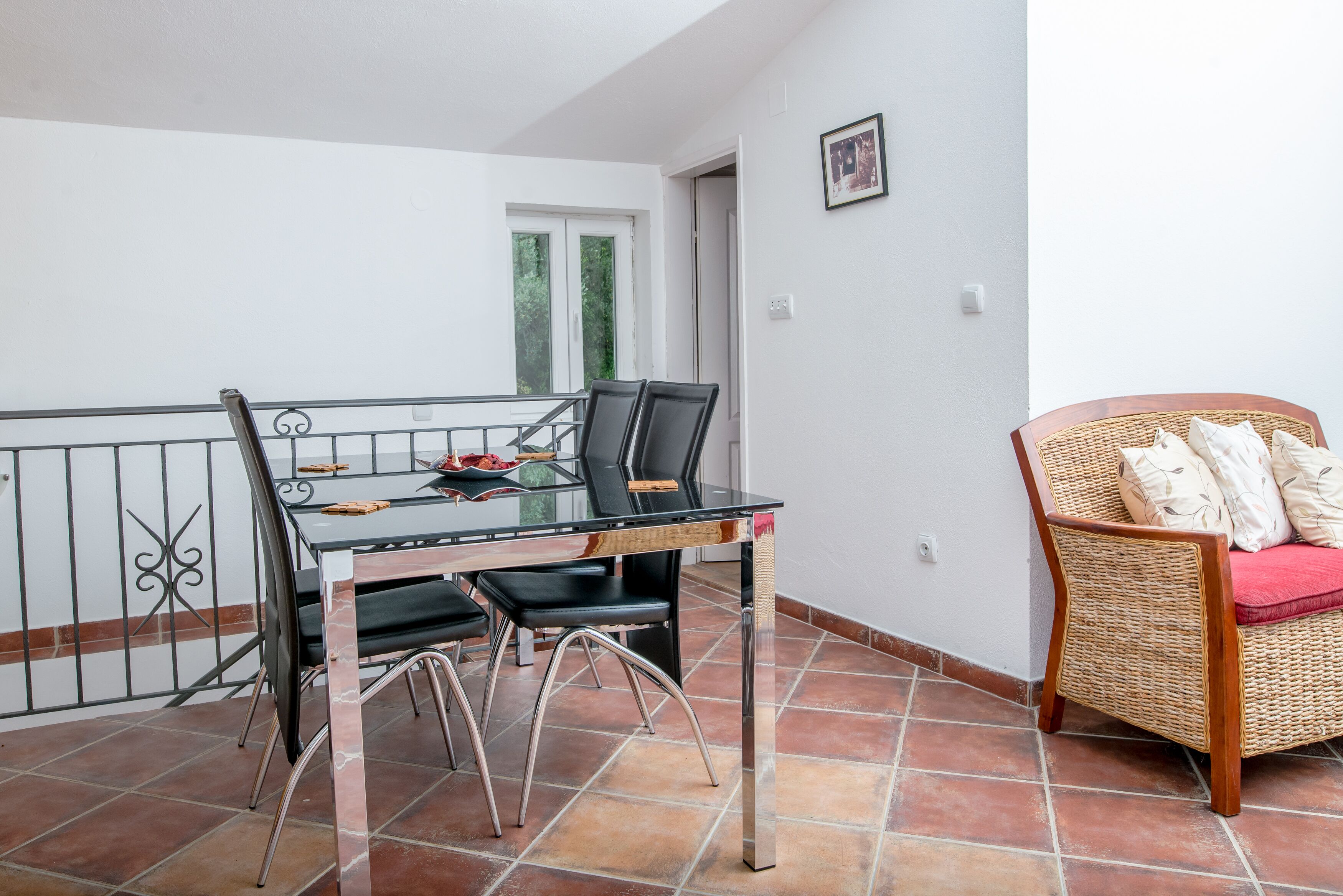 A dining area with a glass table, metal chairs, and a wicker chair with cushions.