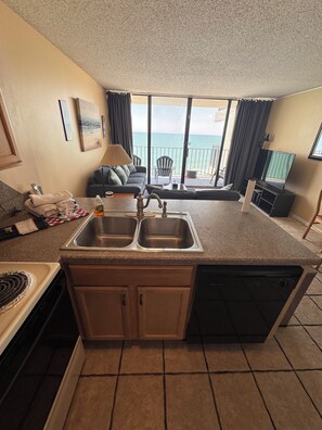 A kitchen with a double sink, a stove, and a dishwasher. There is a dining area with a table and chairs, and a balcony with a view of the ocean.