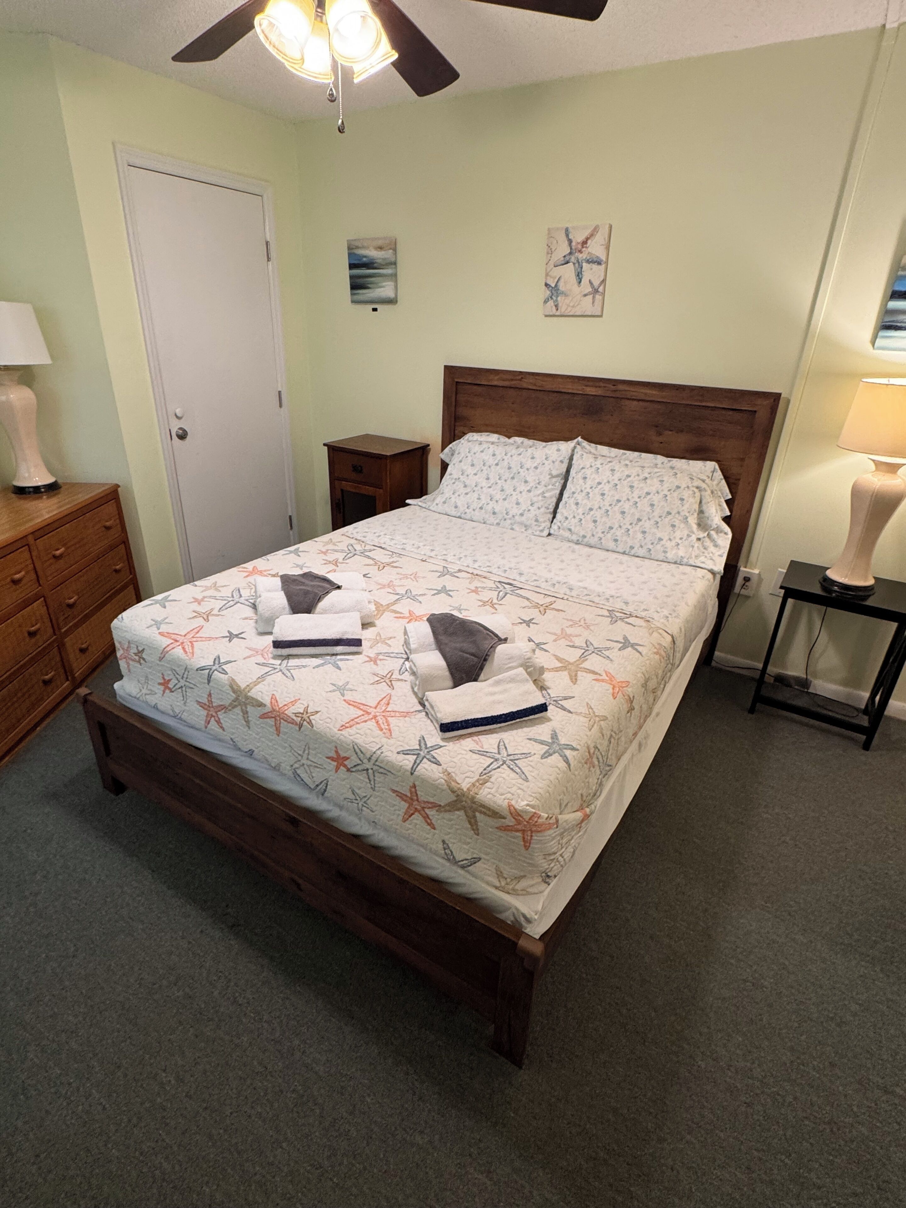 A bedroom with a bed, a nightstand, a dresser, a ceiling fan, and a door.