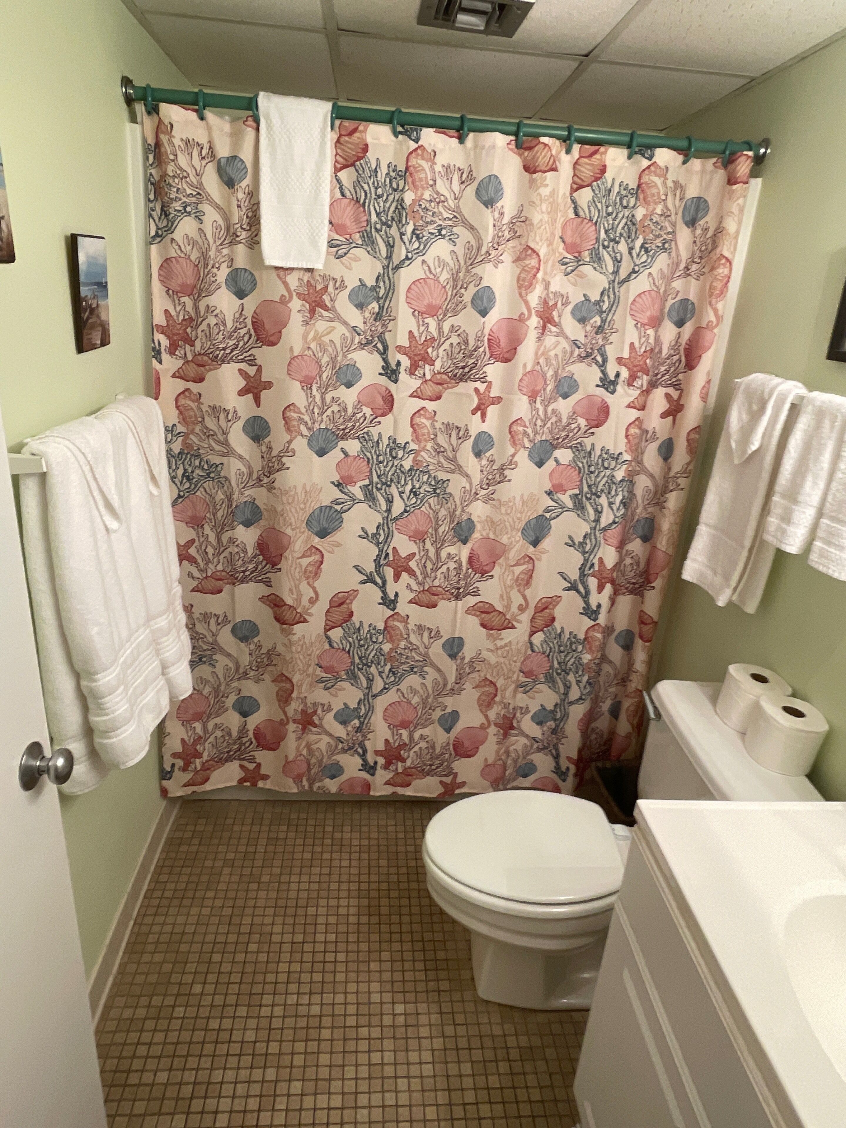 Un baño con suelo de baldosas, un inodoro, una cortina de ducha con estampado floral y un perchero con toallas.