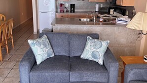 A living room with a gray sofa, a coffee table, and a kitchen area with wooden cabinets, a refrigerator, and a stove. - Family Friendly Fully Equipped two bedrooms two bathrooms Condo in garden city (Murrells Inlet)