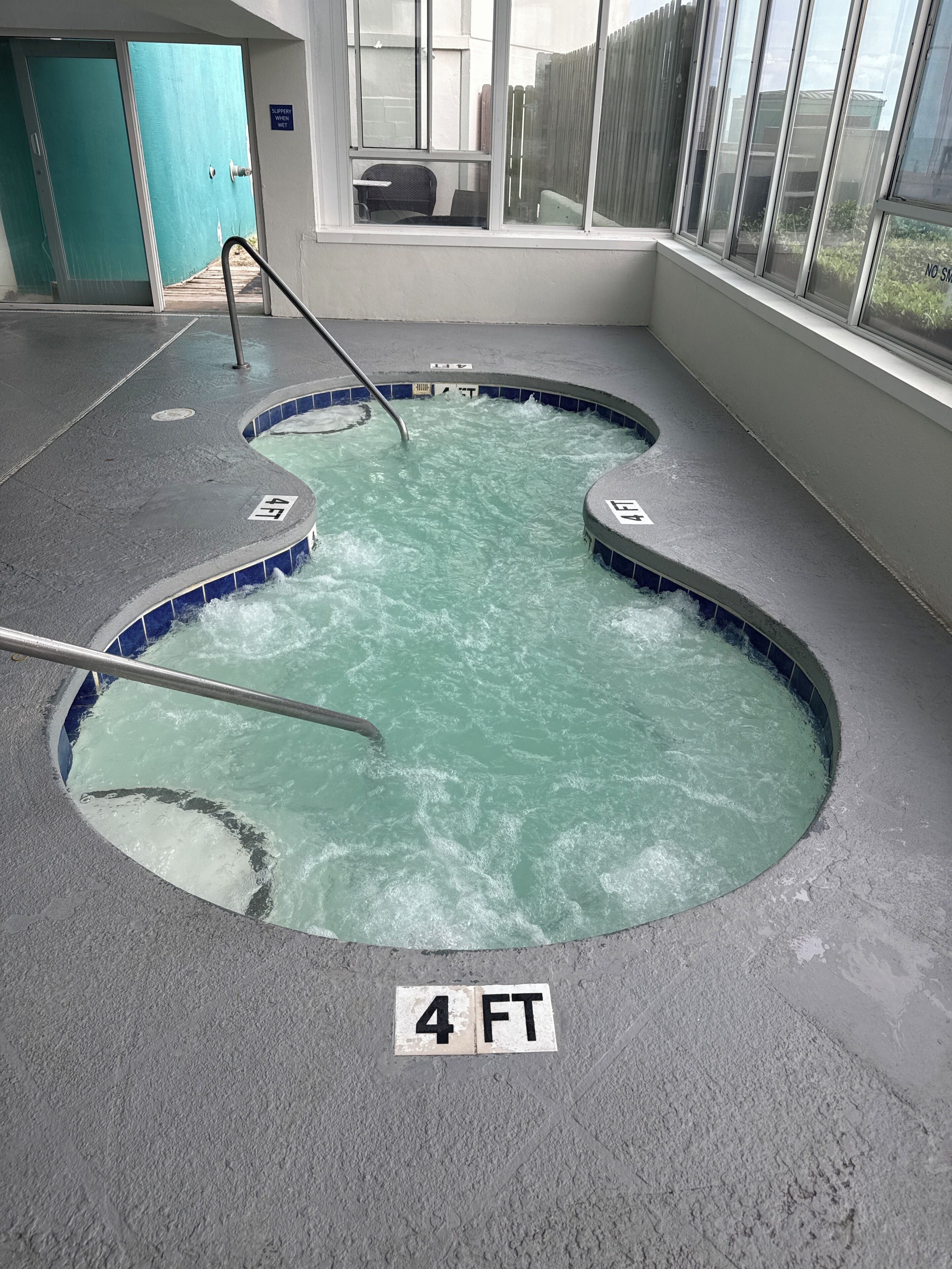 Een jacuzzi met een dieptemarkering van 1,2 meter, omringd door een betegelde vloer en een raam met uitzicht op een hek en gebouwen.