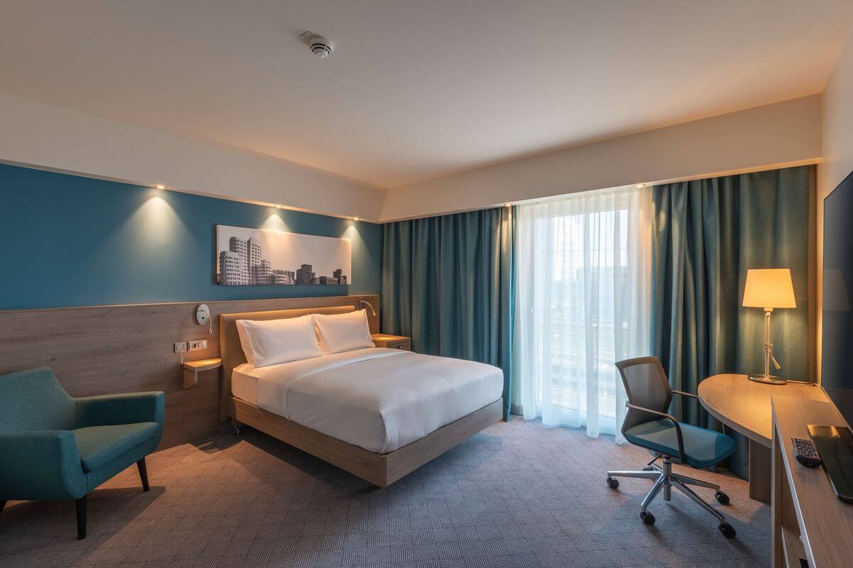 A hotel room with a bed, a desk, a chair, a lamp, and a cityscape artwork on the wall.