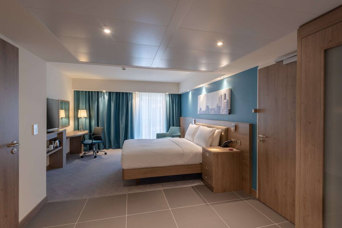 A modern hotel room with a large bed, a desk with a chair, a TV, and a wardrobe.