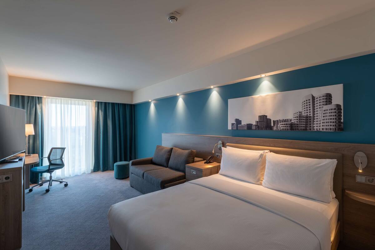 A hotel room with a large bed, a desk with a chair, a sofa, and a cityscape painting on the wall.