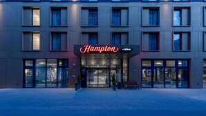 Ingresso moderno di un hotel con insegna al neon, ampie porte in vetro e vialetto lastricato.