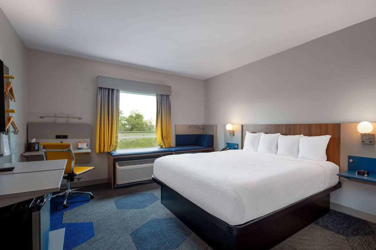 Ein Hotelzimmer mit einem großen Bett, einem Schreibtisch mit Stuhl, einem Fenster mit Vorhängen und einem Nachttisch mit Lampe.