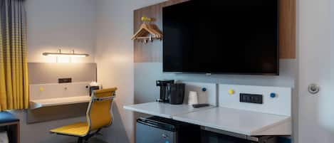 Habitación de hotel con escritorio, silla, microondas, cafetera y televisor montado en la pared.