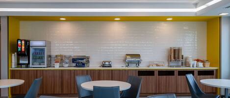 A modern cafe with a yellow accent wall, a coffee machine, a refrigerator, and several seating areas.