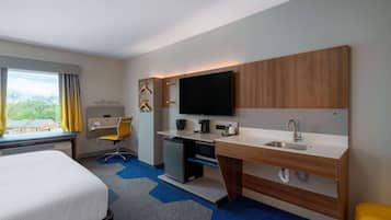 A hotel room with a bed, desk, kitchenette, and a large window with curtains.