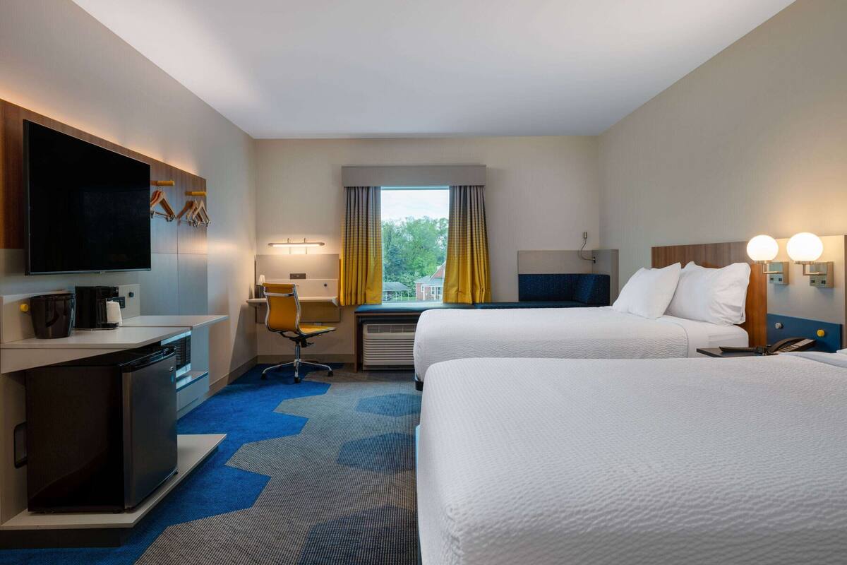 Ein Hotelzimmer mit zwei Betten, einem Schreibtisch mit Stuhl, einem Fernseher, einer Kaffeemaschine und einem Fenster mit Vorhängen.