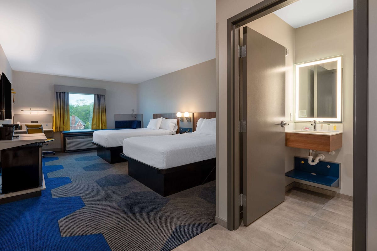 Ein Hotelzimmer mit zwei Betten, einem Schreibtisch, einem Stuhl, einer Schreibtischlampe, einem Fenster mit Vorhängen sowie einem Badezimmer mit Spiegel und Waschbecken.
