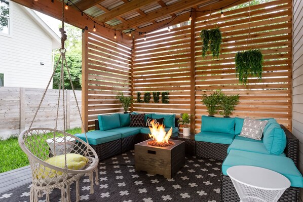 A cozy outdoor seating area with a fire pit, a hanging chair, and a sofa under a wooden slatted roof.