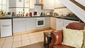 A modern kitchen with white cabinets, a built-in oven, and a dishwasher. There is a wooden table with a magazine and a brown leather chair with a patterned cushion. - Coln Cottage (Cheltenham)