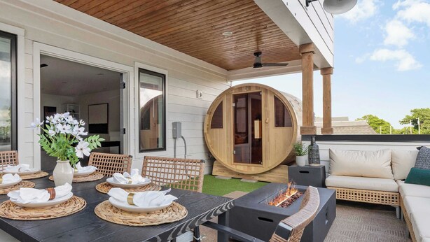 Un área de comedor al aire libre con una mesa negra para cuatro personas, rodeada de sillas de mimbre. Se observa una sauna de madera, un hoyo de fuego y un sofá blanco con cojines.