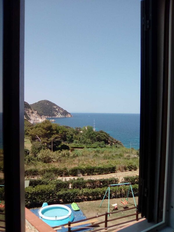 A view from a window overlooking a swimming pool, a grassy area, and a coastal landscape with a hill and the sea.