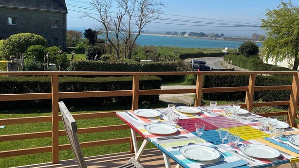 Ein Essbereich im Freien mit Blick auf das Meer, einer Holzterrasse und einem Steinhaus im Hintergrund.