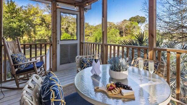 Una terraza de madera con una mesa de vidrio preparada para una comida, rodeada de sillas y vista a árboles.