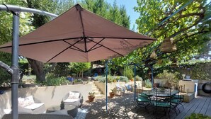 Un patio al aire libre con una mesa y sillas, rodeado de plantas en macetas y una pérgola con una sombrilla marrón.