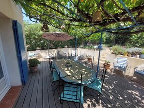 Une terrasse couverte avec une table ronde et des chaises, entourée de plantes en pot et agrémentée d’une pergola avec des jardinières suspendues.