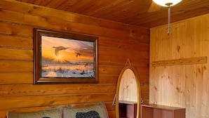 A wooden cabin interior with a bed, a ceiling fan, a framed painting, a bear-themed pillow, and a quilt. - Beautiful country views and wildlife. Less than 2 miles from downtown. (Point Pleasant)