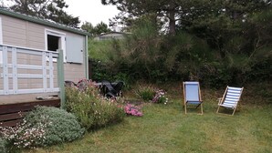 Una cabaña de madera pequeña con un porche, rodeada de un jardín con diversas plantas y flores.
