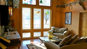 Un salón acogedor con sofá, mesa de centro y chimenea. Hay ventanas amplias con vidrieras, una zona de cocina con fregadero y una puerta que da al exterior.