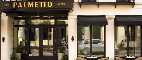 Exterior del Hotel Palmetto con un toldo negro, grandes puertas de vidrio y zona de estar al aire libre con sillas y mesas.