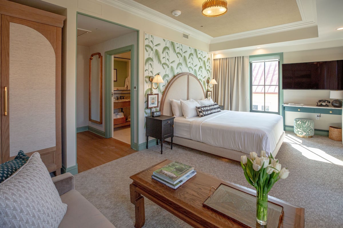 Habitación de hotel con cama, escritorio, televisor, mesa de centro y un jarrón de flores.
