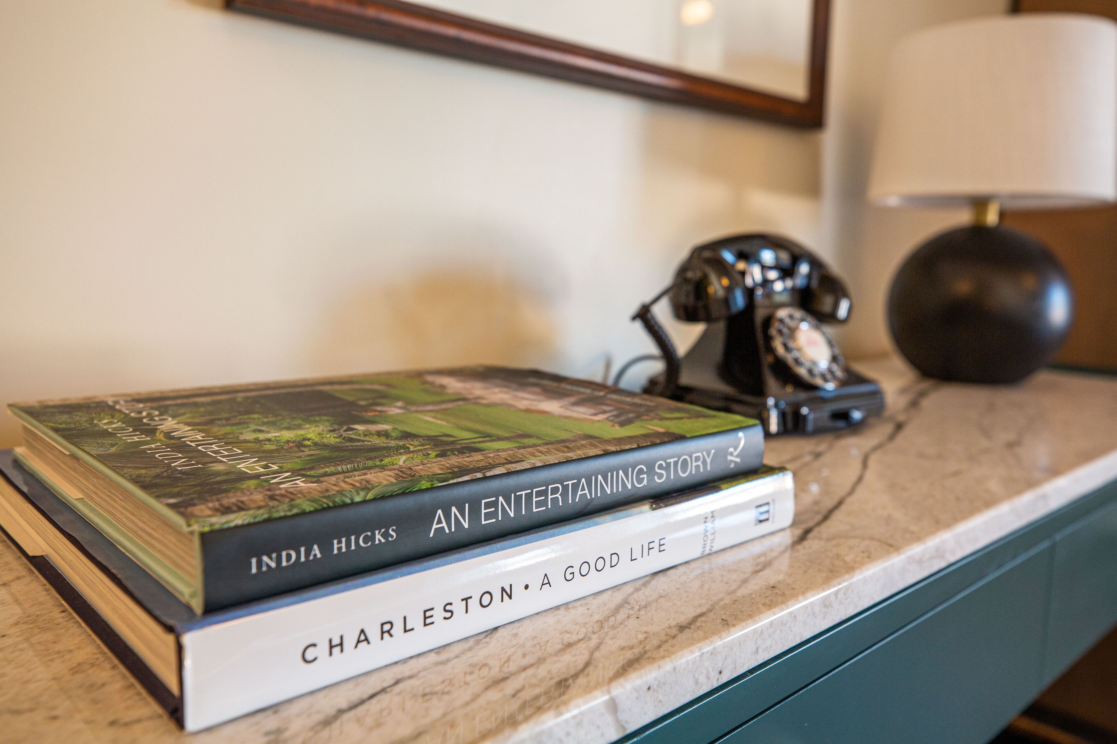 A stack of books on a marble countertop with a vintage telephone and a lamp in the background.