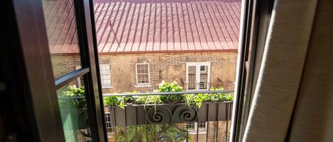 A view through a window with a metal balcony railing, a brick building with a red roof, and some greenery.