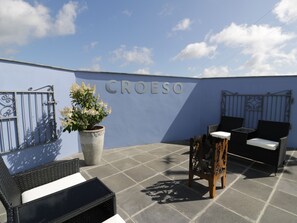 A rooftop patio with a blue wall, a potted plant, wicker furniture, and a metal gate. - Drws y Gorlan (Llanerchymedd)