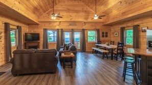 A spacious cabin interior with a wooden ceiling, a fireplace, a sofa, a dining table, and a kitchen area. - Incredible Views, Hot Tub, Pool Table, Waterfalls, Hiking (Bryson City)