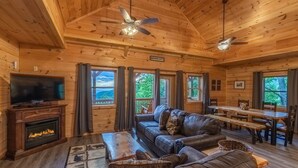A cozy cabin interior with a fireplace, leather sofa, dining table, and large windows offering a view of the outdoors. - Incredible Views, Hot Tub, Pool Table, Waterfalls, Hiking (Bryson City)