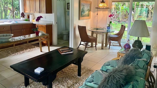 A cozy living room with a sofa, a coffee table, and a dining area with chairs and a table. There is a kitchen visible in the background. - JJs Hāna Hale - Farm House (Hana)