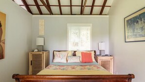 A bedroom with a wooden bed, two bedside tables with lamps, a window with blinds, and a painting on the wall. - JJs Hāna Hale - Farm House (Hana)