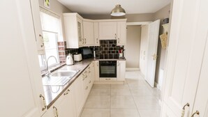 A kitchen with white cabinets, a black tiled backsplash, a built-in oven, and a window with a white curtain. - Curragh Hill (Mulranny)