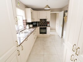 Una cocina con armarios blancos, salpicadero de azulejos negros, horno empotrado y ventana con cortina blanca.